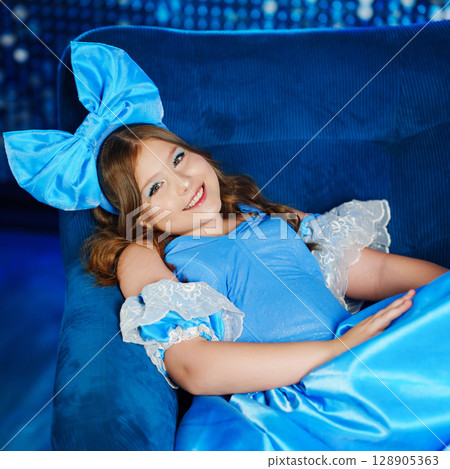Cute teenage girl in blue doll dress with big blue bow posing in an armchair  128905363