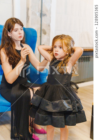 Mom gathers hair for a beautiful little daughter in a smart black dress. 128905365