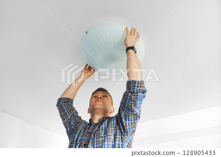 A man in a plaid shirt holds a large white round ceiling lamp in his hands. 128905433