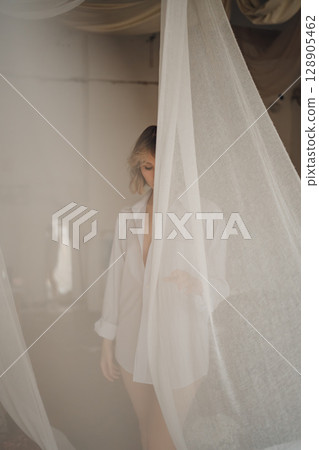 An attractive woman in a white shirt on a nude barely in tulle from the window. An attractive woman in a white shirt on a nude barely in tulle from the window. 128905462