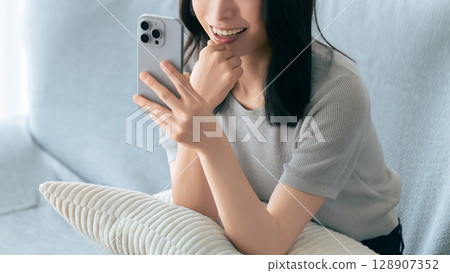 Smiling woman sitting on sofa and using smartphone Smiling woman sitting on sofa and using smartphone 128907352