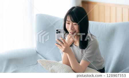 Smiling woman sitting on sofa and using smartphone Smiling woman sitting on sofa and using smartphone 128907353