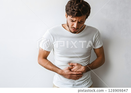 A man stands against a plain background, experiencing abdominal pain, clutching his stomach with both hands, showing clear signs of distress. 128909847