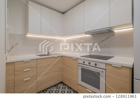 Modern kitchen with white cabinets and wooden accents. Features a marble backsplash, stainless steel appliances, and patterned floor tiles. 128911961