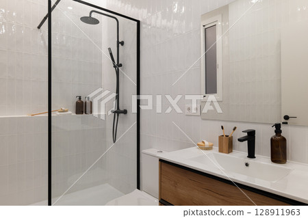 Modern bathroom with a glass shower, white tiles, wooden vanity, and minimalistic decor. Natural light enters through a window. Modern bathroom with a glass shower, white tiles, wooden vanity, and minimalistic decor. Natural light enters through a window. 128911963