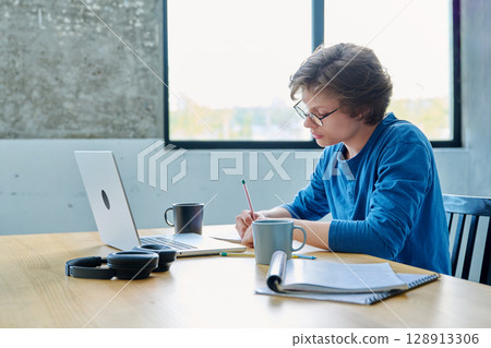 Teenage guy student sitting at the table with laptop, textbooks in home interior 128913306