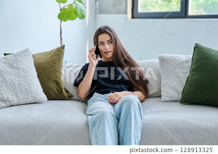 Young woman sitting at home on couch talking on smartphone Young woman sitting at home on couch talking on smartphone 128913325