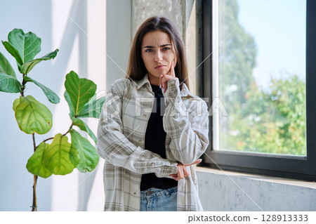 Portrait of serious teenage girl looking at camera near window 128913333