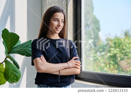 Portrait of confident successful teenage girl posing with crossed arms near window 128913335