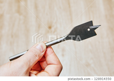 Flat drill bit with sharp cutting edges and small round hole near tip held on neutral background closeup shows woodworking drilling tool. Flat drill bit with sharp cutting edges and small round hole near tip held on neutral background closeup shows woodworking drilling tool. 128914350