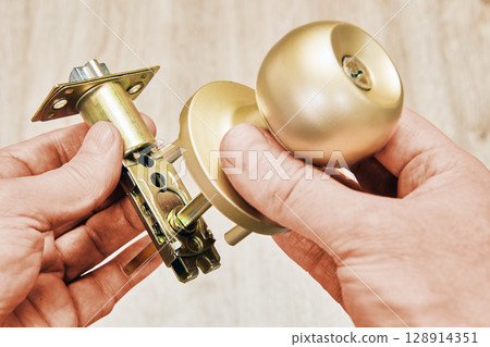Hands holding brass door knob with spring latch mechanism ready for installation on wooden door. Hands holding brass door knob with spring latch mechanism ready for installation on wooden door. 128914351