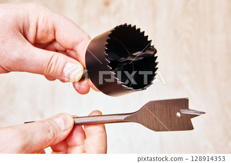 Spade bit and hole saw drill bit for woodworking held in hands close to each other ready to be attached to cordless drill for woodwork tasks. Spade bit and hole saw drill bit for woodworking held in hands close to each other ready to be attached to cordless drill for woodwork tasks. 128914353