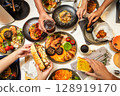 Captured from a bird's-eye view, five or six people are dining together at a Western-style restaurant, enjoying spaghetti, croissants, chicken wings, coffee, and other dishes. 128919170