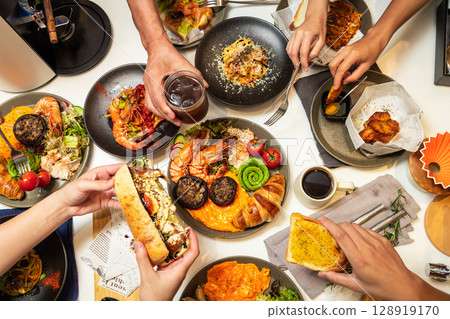 Captured from a bird's-eye view, five or six people are dining together at a Western-style restaurant, enjoying spaghetti, croissants, chicken wings, coffee, and other dishes. Captured from a bird's-eye view, five or six people are dining together at a Western-style restaurant, enjoying spaghetti, croissants, chicken wings, coffee, and other dishes. 128919170
