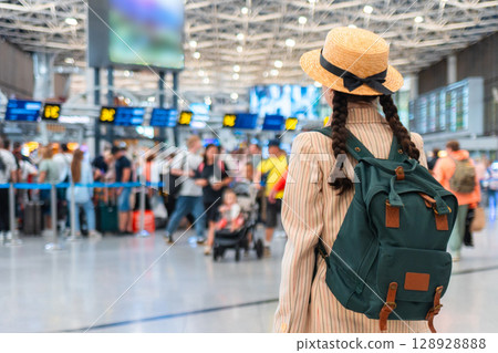 Back view of woman in an elegant suit, straw hat and backpack in airport. Copy space. A blurred departure lounge in the background. Concept of trip and travel Back view of woman in an elegant suit, straw hat and backpack in airport. Copy space. A blurred departure lounge in the background. Concept of trip and travel 128928888
