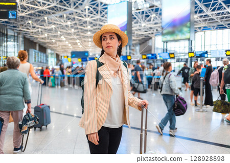 Pretty Caucasian young woman in straw hat wheeling a suitcase to the check-in desk. Defocused people in background. The concept of vacation and flight trip Pretty Caucasian young woman in straw hat wheeling a suitcase to the check-in desk. Defocused people in background. The concept of vacation and flight trip 128928898