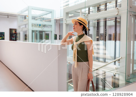 Mid shot of young pretty Caucasian elegant woman in suit and straw hat holds bag and posing in the modern art gallery. Concept of cultural education and Museum day 128928904