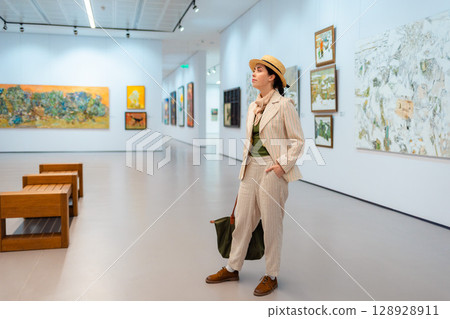 Museum day. Wide shot of young Caucasian pretty woman in elegant suit and straw hat posing in the art gallery. Concept of cultural education Museum day. Wide shot of young Caucasian pretty woman in elegant suit and straw hat posing in the art gallery. Concept of cultural education 128928911