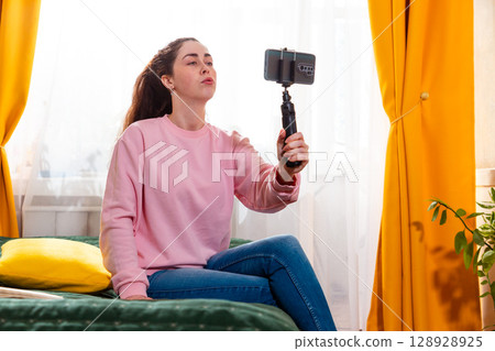 Mid shot of young woman recording an inspiring vlog for social media while sitting on a bed, using a smartphone on a tripod. 128928925