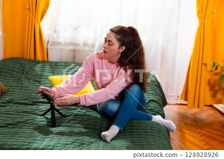 Mid shot of young Caucasian woman recording a social media vlog while sitting on a bed, discussing personal branding. 128928926