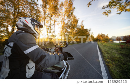 Driver riding motorcycle on empty road during sunset , view from the passenger side Driver riding motorcycle on empty road during sunset , view from the passenger side 128929213