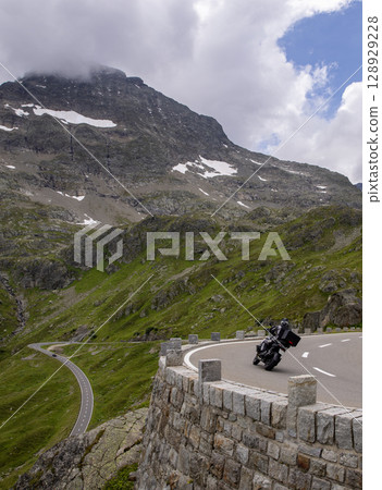 An alpine road with a sharp curve and motorcycle driver. Beautiful view of the high mountain pass. 128929228