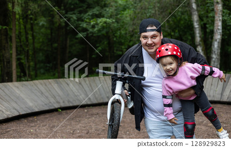 A father holds his smiling daughter in protective gear after riding a balance bike in the park, promoting family bonding and outdoor activity. Happy father holding his cute daughter in his arms. 128929823