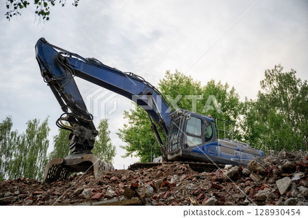 Blue excavator working on demolition site with pile of broken bricks and concrete rubble 128930454