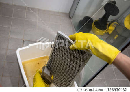 Person cleaning greasy metal range hood filter with sponge and rubber gloves over dirty water 128930482