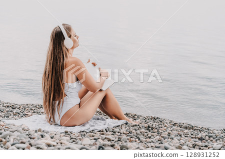 A woman is sitting on the beach with a laptop and headphones on. She is wearing a white bikini and she is enjoying her time by the water. A woman is sitting on the beach with a laptop and headphones on. She is wearing a white bikini and she is enjoying her time by the water. 128931252