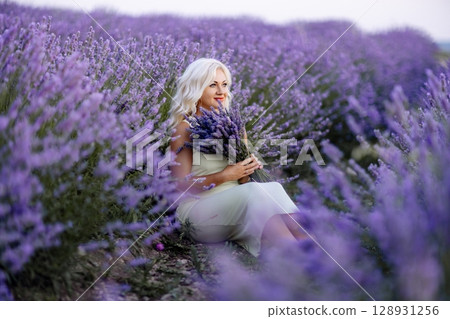 Blonde woman poses in lavender field at sunset. Happy woman in white dress holds lavender bouquet. Aromatherapy concept, lavender oil, photo session in lavender 128931256