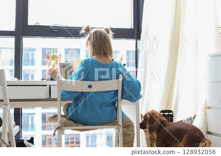 young girl seating at the table in your room near windows 128932056