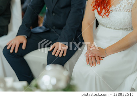 Close-up of bride and groom sitting together during wedding, showing joined hands and elegant attire Close-up of bride and groom sitting together during wedding, showing joined hands and elegant attire 128932515