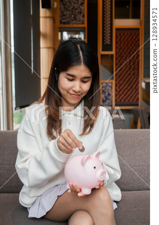 A young woman in a cozy living room saves money by placing a coin into a pink piggy bank, symbolizing financial planning and savings. 128934571