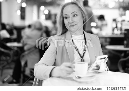 Woman sits at table in restaurant, drinks coffee and writes a message on phone. Woman sits at table in restaurant, drinks coffee and writes a message on phone. 128935785