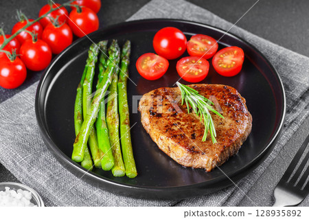 Grilled beef steak closeup with asparagus and cherry tomatoes on black plate, gourmet healthy dinner Grilled beef steak closeup with asparagus and cherry tomatoes on black plate, gourmet healthy dinner 128935892