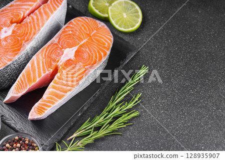 Fresh raw salmon or trout steaks with tomatoes, spices, rosemary and lime on black stone background Fresh raw salmon or trout steaks with tomatoes, spices, rosemary and lime on black stone background 128935907