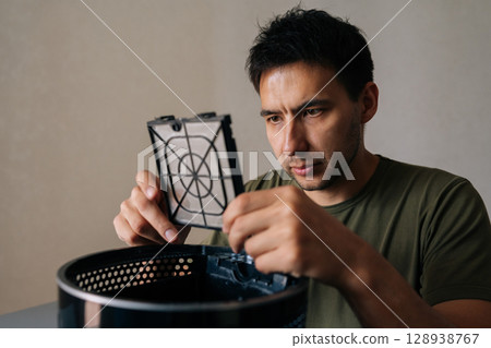 Close-up face of focused male checking air humidifier filter, carefully examining device component for maintaining indoor air quality and home wellness. Concept of maintenance modern home appliance. Close-up face of focused male checking air humidifier filter, carefully examining device component for maintaining indoor air quality and home wellness. Concept of maintenance modern home appliance. 128938767
