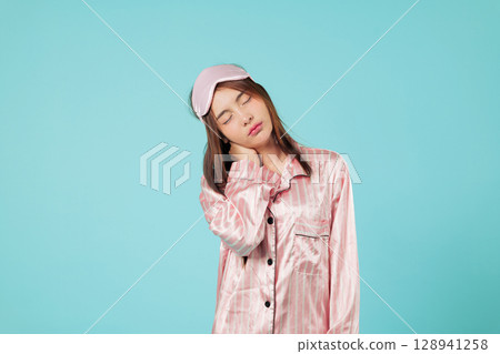Asian woman experiences neck and shoulder pain, feeling tired needing sleep, wearing pajamas and sleep mask, possibly stretching after long sleep, headshots isolated studio blue background Asian woman experiences neck and shoulder pain, feeling tired needing sleep, wearing pajamas and sleep mask, possibly stretching after long sleep, headshots isolated studio blue background 128941258