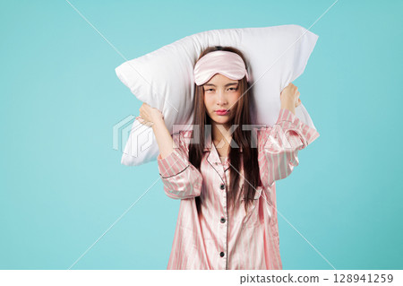 Stressed Asian woman with pillow covers her ears because of snore, pajama and sleep mask show her bad mood, needs sleep as she tries to rest at home tonight, studio isolated blue background Stressed Asian woman with pillow covers her ears because of snore, pajama and sleep mask show her bad mood, needs sleep as she tries to rest at home tonight, studio isolated blue background 128941259