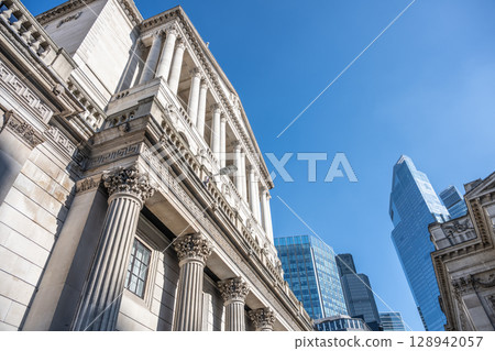 The historic Bank of England building stands prominently against a clear blue sky. Modern skyscrapers surround this iconic structure, showcasing London's architectural blend of old and new. 128942057