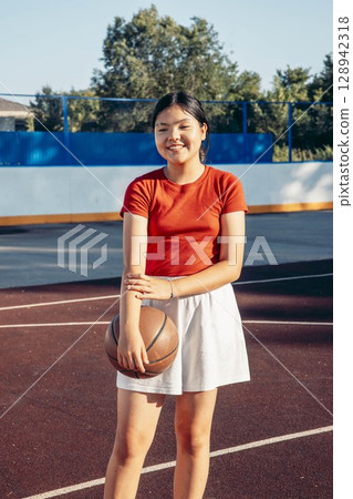 smiling Asian teenage girl in red shirt, white skirt holding basketball on outdoor court on sunny summer day, youth sport girl empowerment healthy lifestyle and leisure physical activity 128942318