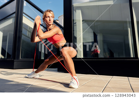 Woman performing resistance band exercise outdoors near a modern glass building Woman performing resistance band exercise outdoors near a modern glass building 128943043