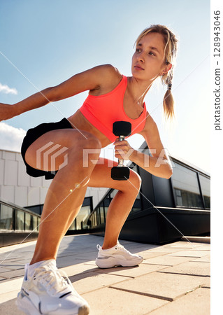 Determined athletic woman training outside with weights on a sunny day Determined athletic woman training outside with weights on a sunny day 128943046