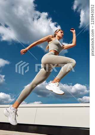 Woman in sportswear leaping outside with a blue sky and clouds background 128943052