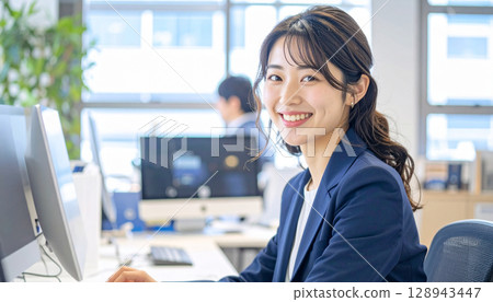A young female office worker smiling and operating a PC in a clean office 128943447