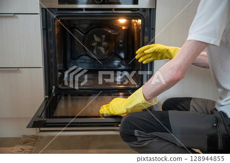 Person cleaning oven door glass with sponge with yellow rubber gloves 128944855