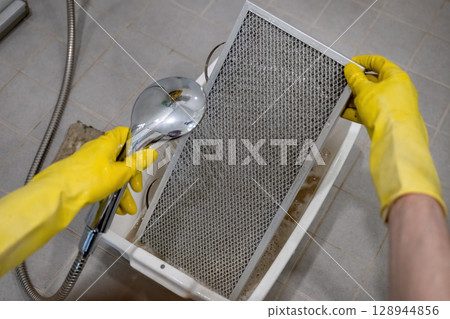 Person rinsing metal kitchen range hood filter with water in sink with yellow rubber gloves 128944856