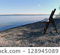 A wooden folding chair with a white hat rests on a rocky beach by a calm lake under a clear blue sky. Evokes a sense of tranquility, relaxation, and vacation. Tranquil beach scene 128945089