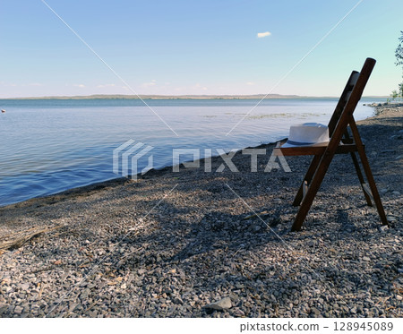 A wooden folding chair with a white hat rests on a rocky beach by a calm lake under a clear blue sky. Evokes a sense of tranquility, relaxation, and vacation. Tranquil beach scene A wooden folding chair with a white hat rests on a rocky beach by a calm lake under a clear blue sky. Evokes a sense of tranquility, relaxation, and vacation. Tranquil beach scene 128945089